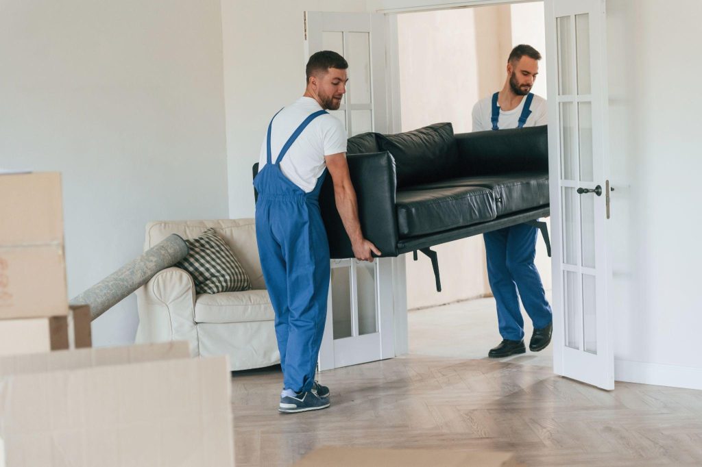 Two men in blue overalls carry a black sofa through a white door in a bright room. Cardboard boxes and a rolled carpet are in the background.
